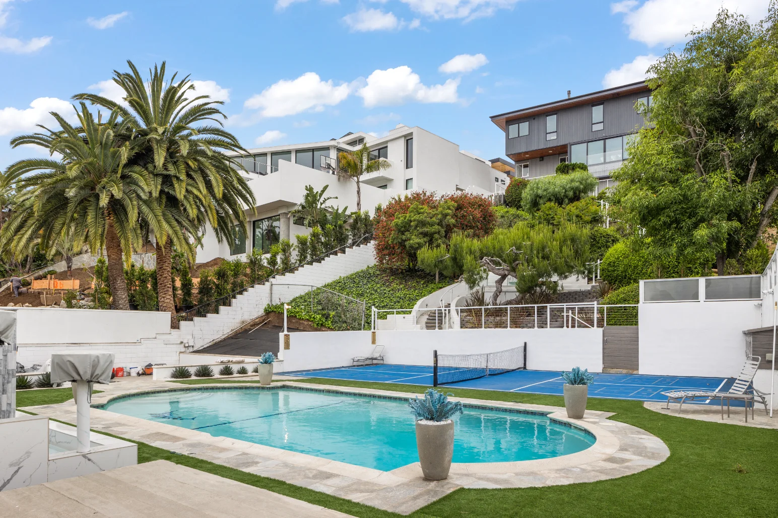 Modern backyard featuring a swimming pool, lounge chairs, potted plants, an inviting pickleball or tennis court, palm tree, and hillside homes. Thoughtfully designed outdoor spaces create the perfect setting under a blue sky.
