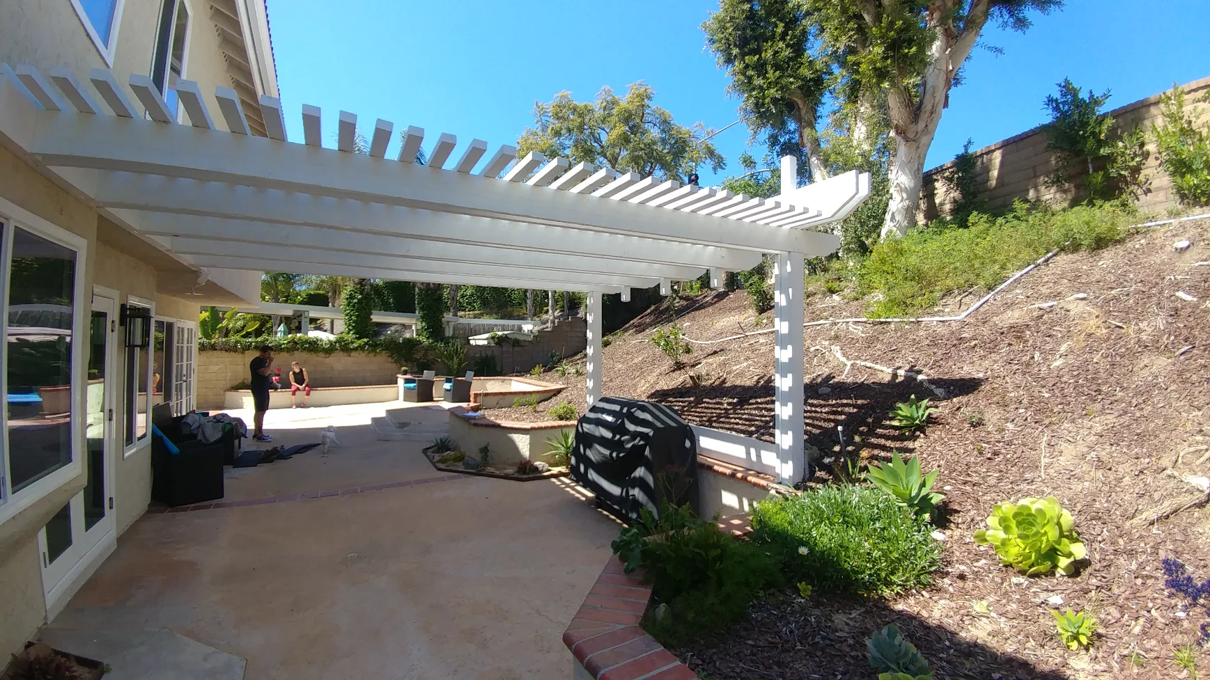 A white pergola covers a patio next to a beige house, with a sloped garden and retaining wall in the background under a clear blue sky.