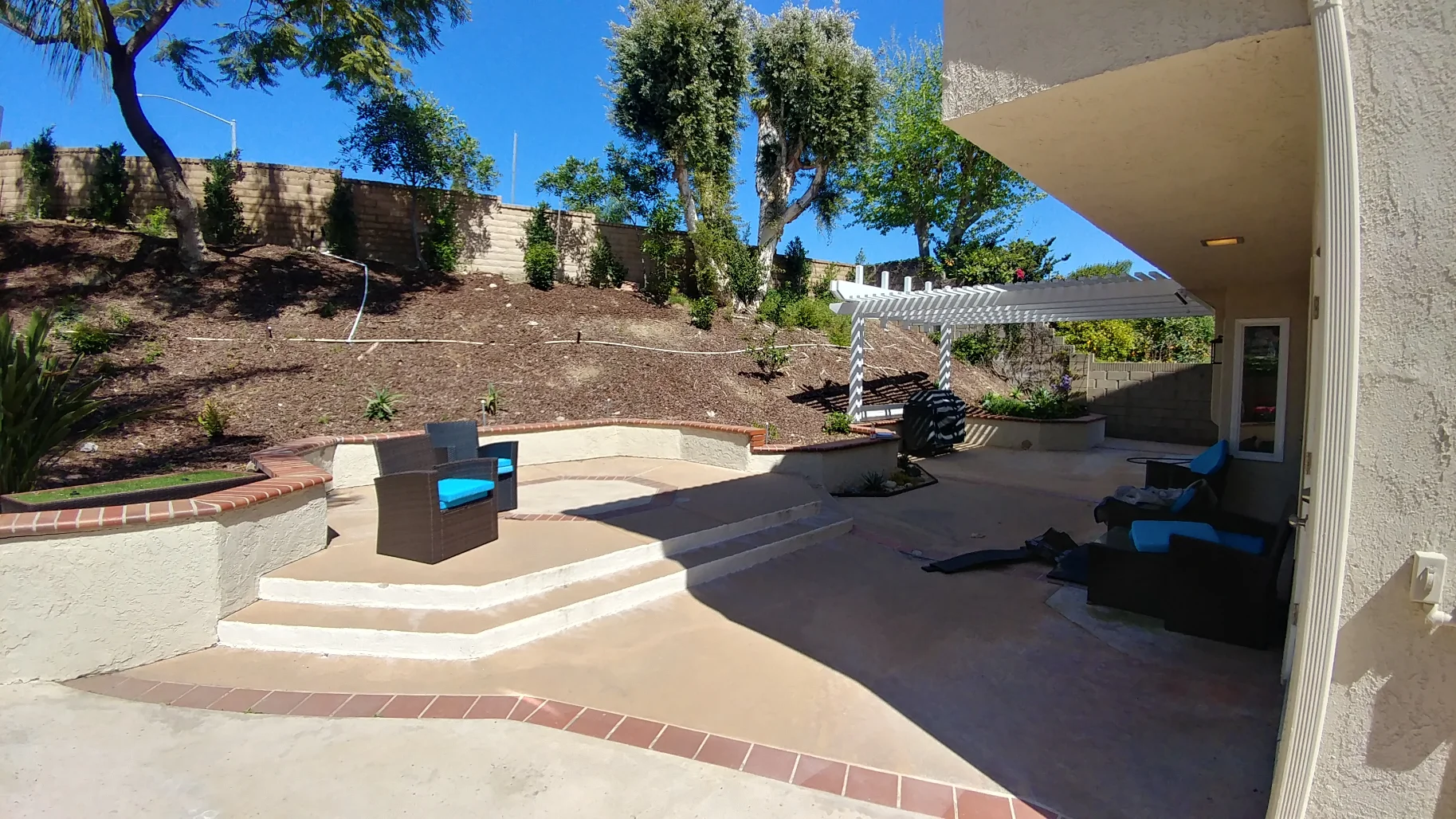 Outdoor patio area with beige seating, blue cushions, a pergola, and a tiered retaining wall with sparse landscaping under a clear blue sky.
