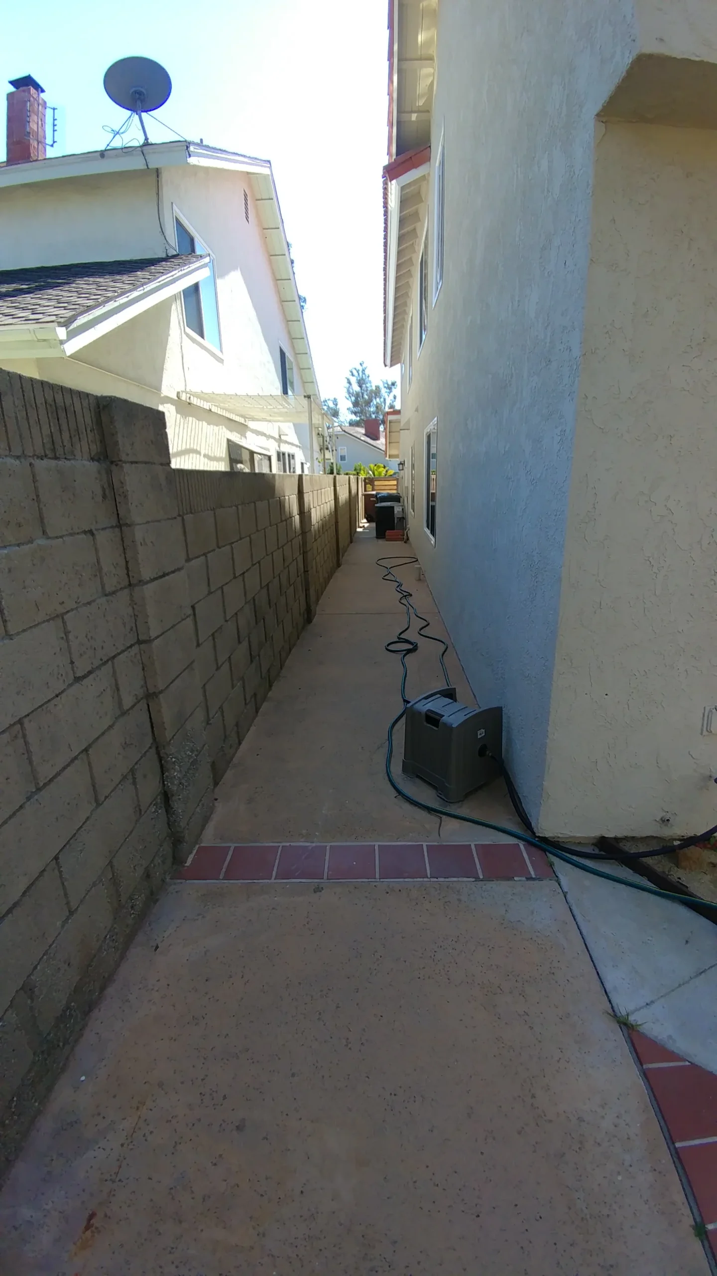 A narrow concrete walkway runs between a block wall and a house, with a hose and equipment placed along the path.