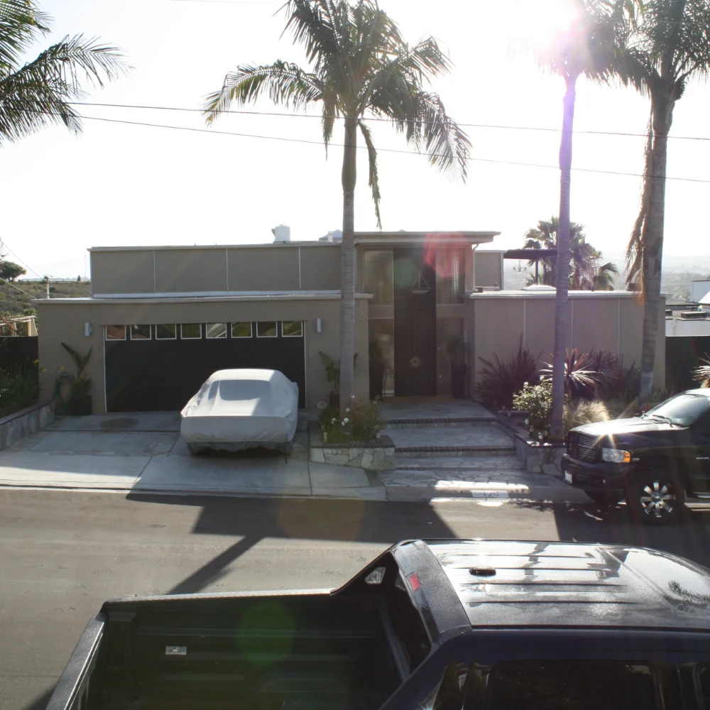 A modern, single-story house with a flat roof and palm trees features inviting outdoor living spaces. A covered car rests in the driveway, while two trucks are parked on the street in front.