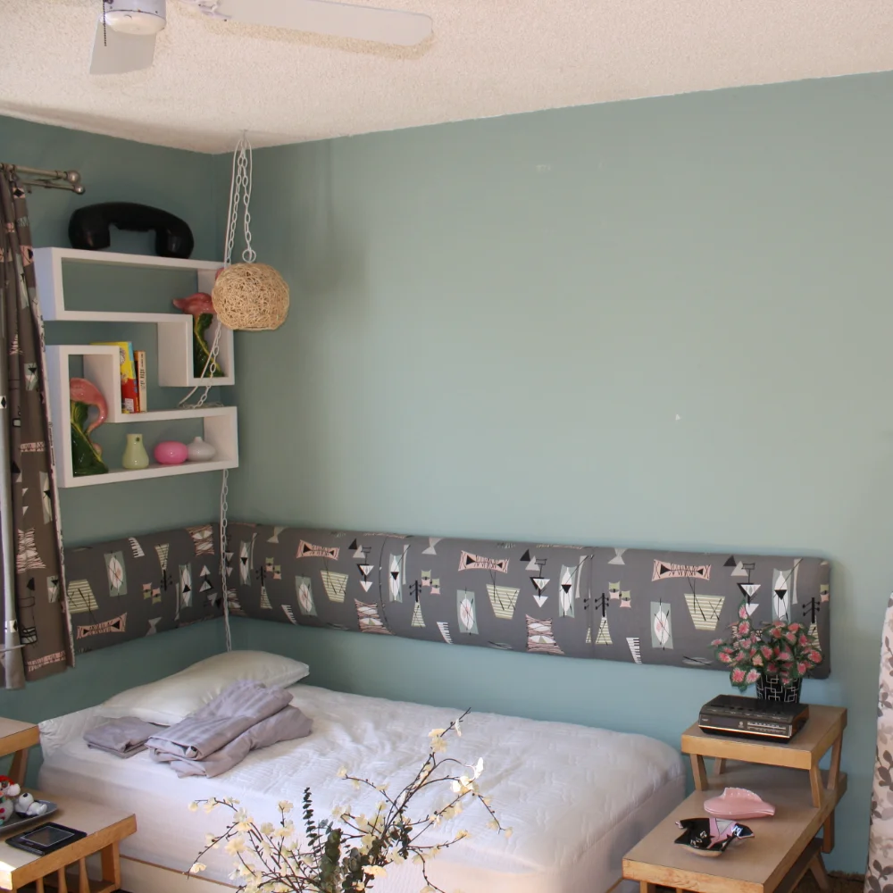 A small bedroom with a single bed, folded towels, two wooden side tables, wall shelves with books and decor, and a light blue accent wall—ideal as a cozy ADU or guest retreat within your whole home remodel.