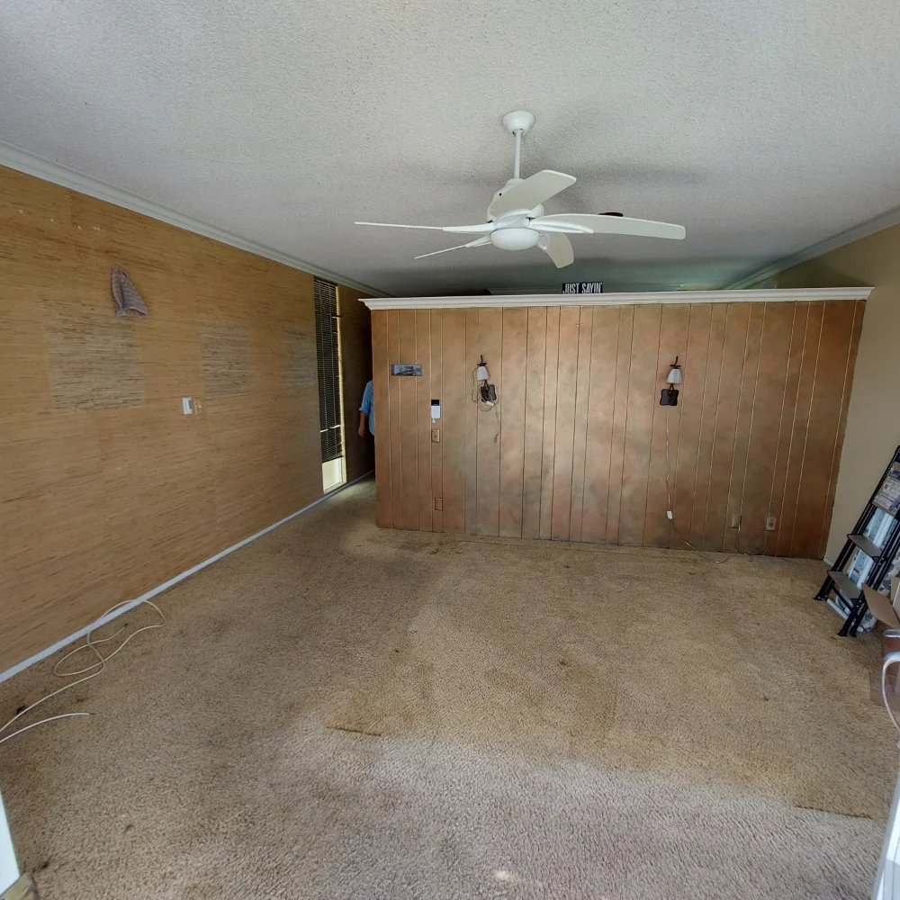 Empty room with brown carpet, wood-paneled partition wall, ceiling fan, exposed cords, and shelves on the right; some wall damage and stains on carpet—great potential for an ADU or whole home remodel.