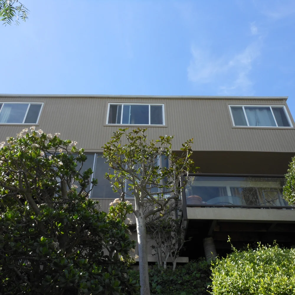 A two-story beige house with large windows, partially obscured by green trees and shrubs, offers inviting outdoor living spaces. The home is viewed from below against a blue sky.