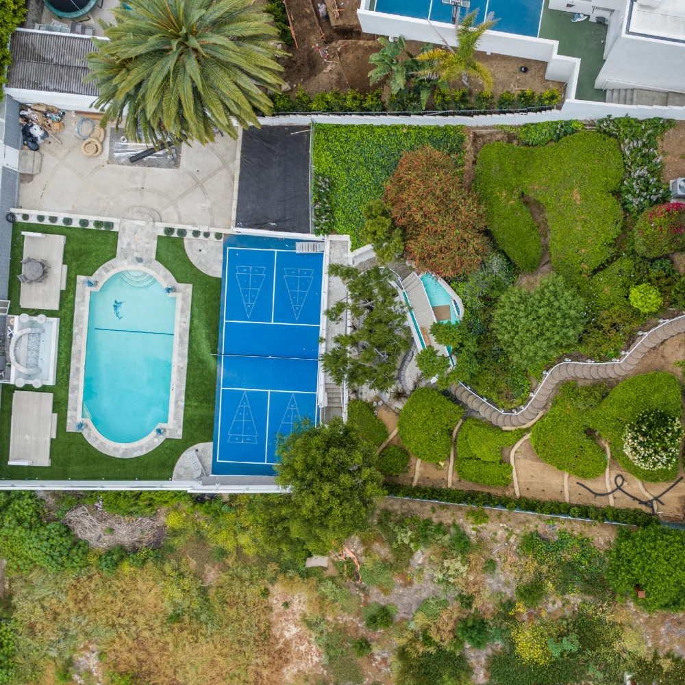 Aerial view of a backyard with a swimming pool, blue pickleball court, landscaped garden, trees, an ADU, and a winding pathway—perfect for outdoor living.