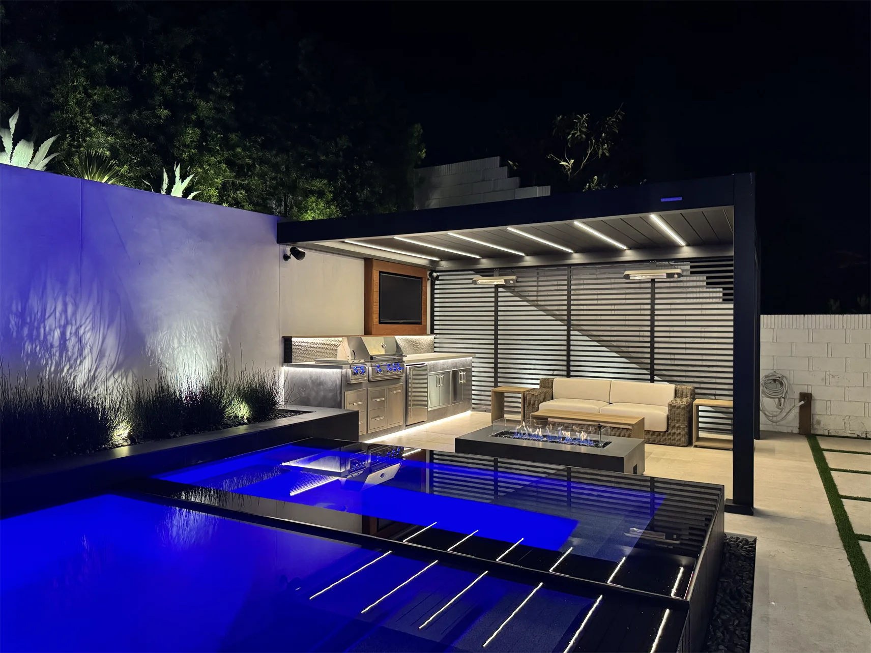 Modern outdoor patio in Laguna Hills at night with illuminated blue pool, stainless steel grill, countertop, TV, and covered seating area featuring a sofa and fire pit—an inspiring home remodel and landscape remodel masterpiece.
