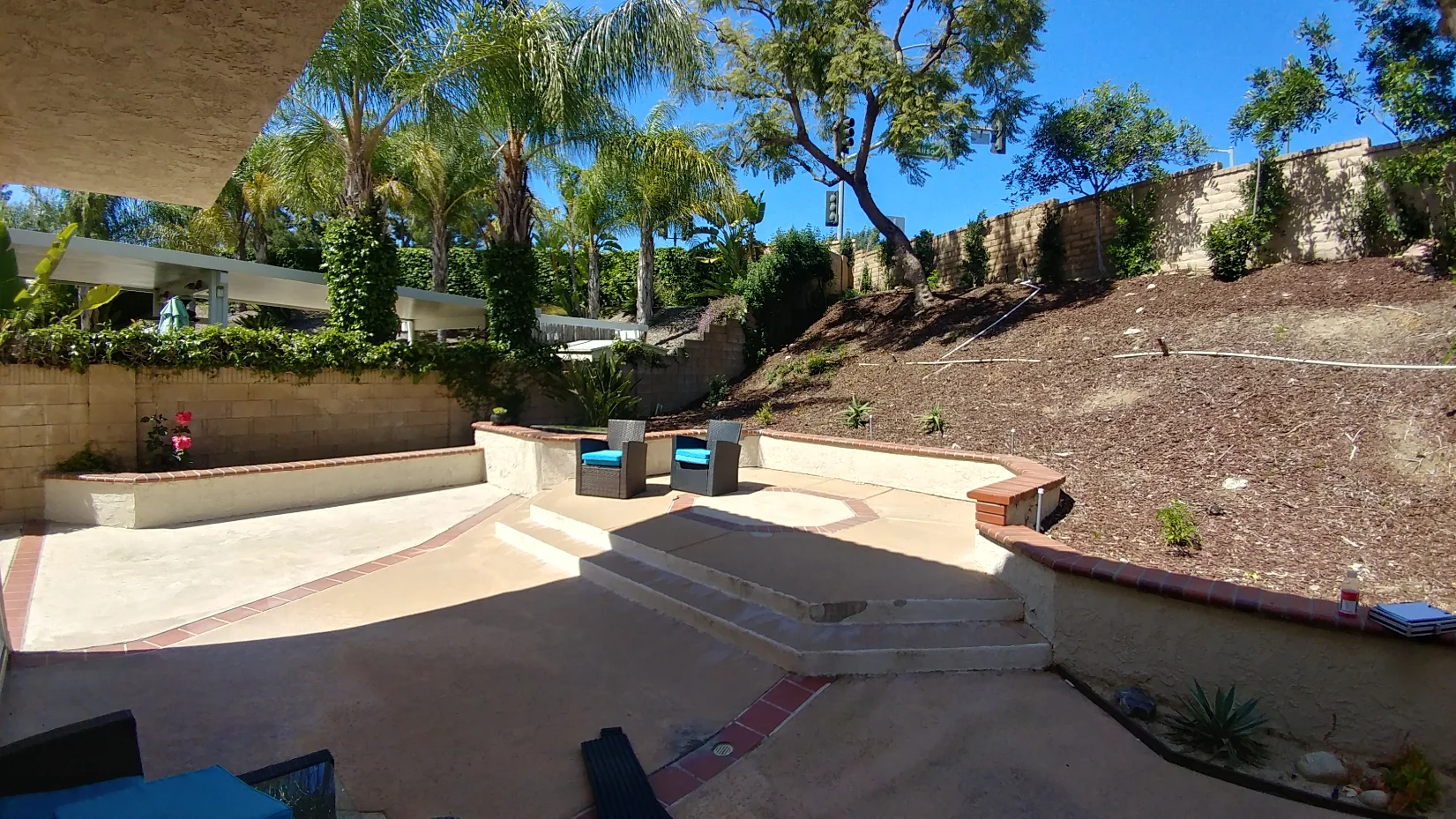 A backyard patio with built-in seating, two blue chairs, steps, and a sloped garden with trees and shrubs under a clear blue sky.