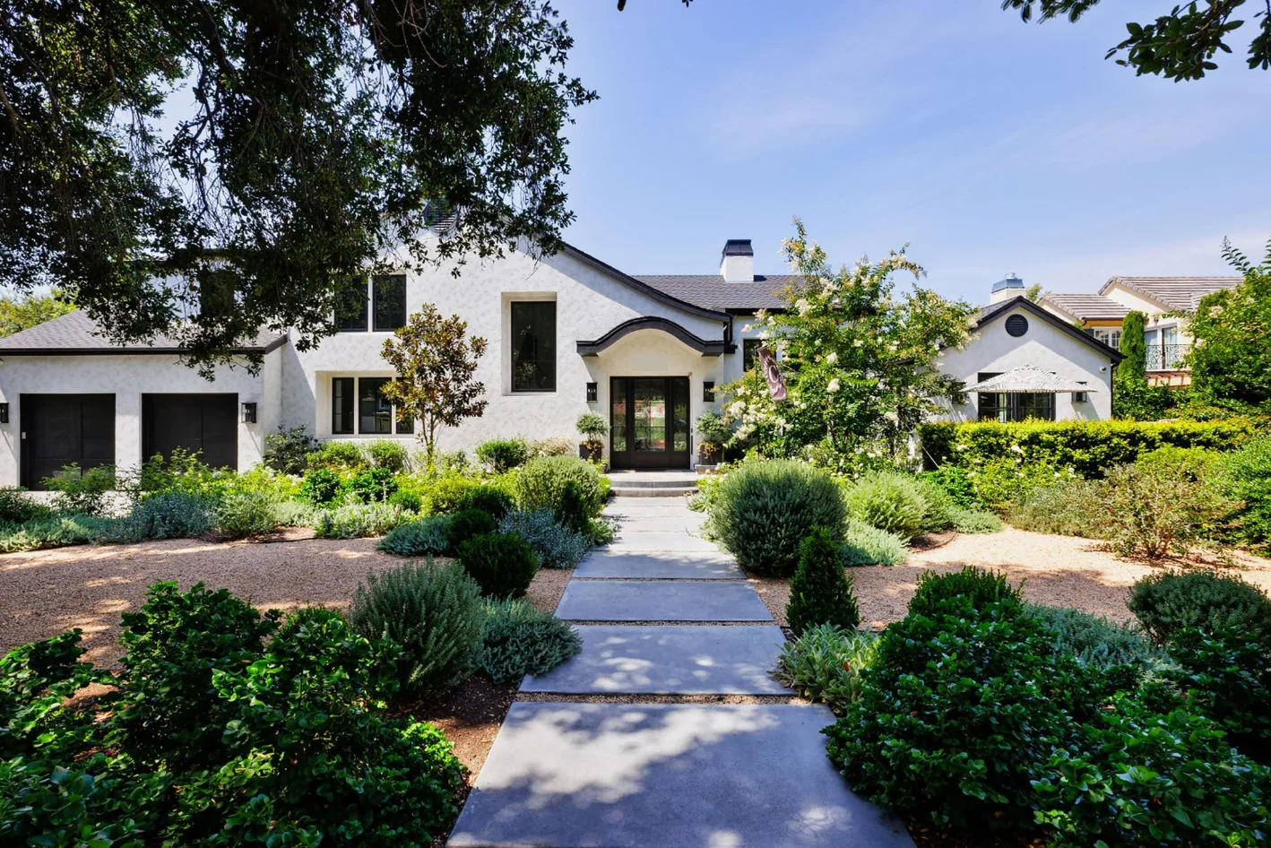 A modern white house with black trim, a Custom Home Build in Santa Ana, CA, is surrounded by green landscaping, shrubs, and trees, with a stone pathway leading to the front entrance.