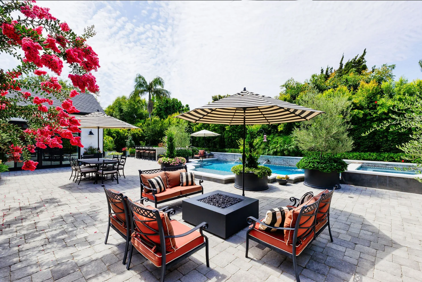 Outdoor patio area with metal seating around a square fire pit, striped umbrellas, dining table, pool, hot tub, and lush greenery—a stunning highlight of this custom home build in Santa Ana, CA.