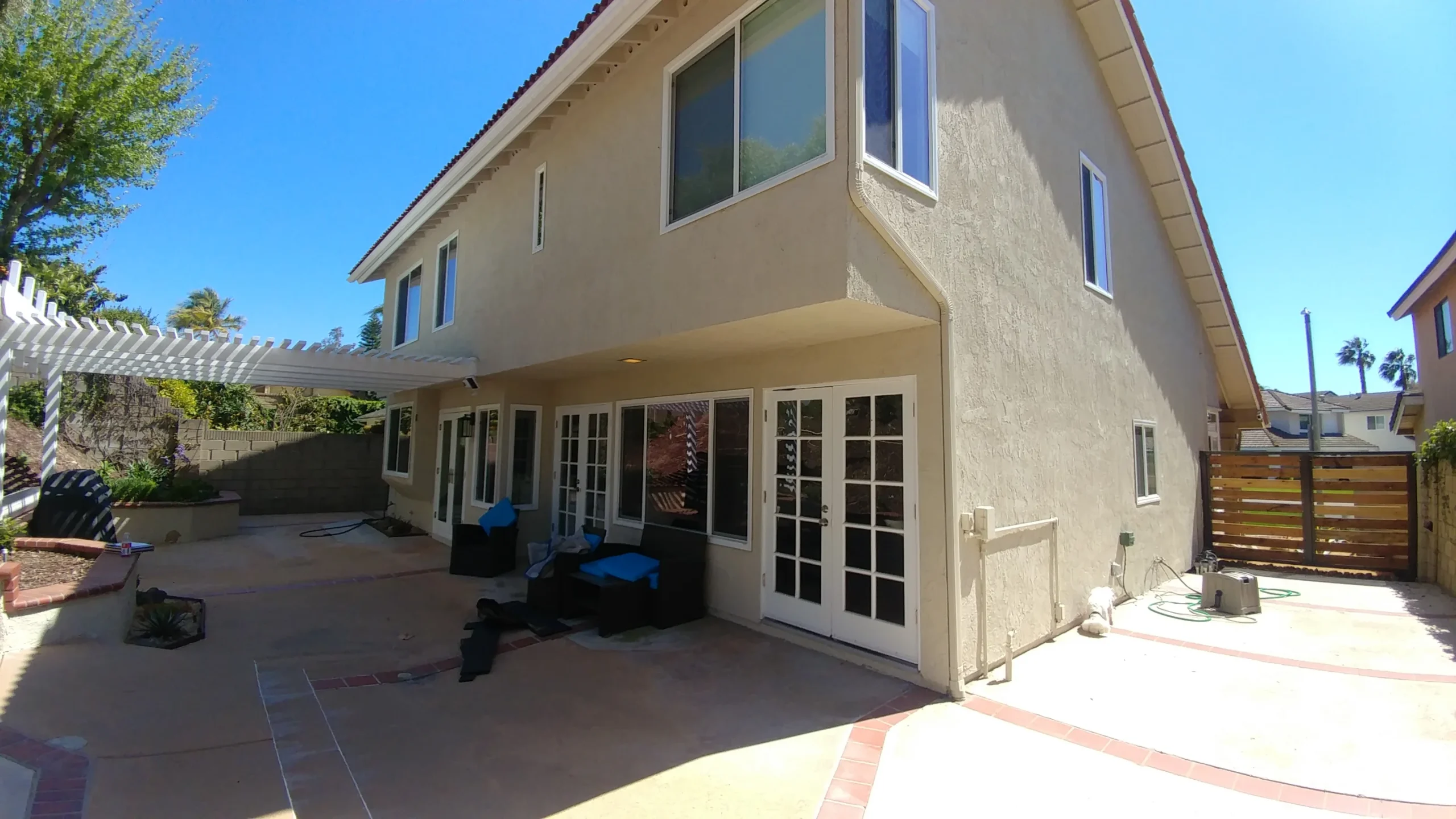 A two-story beige house with large windows and multiple glass doors opens onto a spacious patio with outdoor seating under a white pergola.