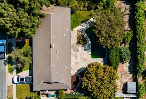 Aerial view of a single-story house with a gray roof, driveway, parked car, green lawn, and surrounding trees and shrubs.