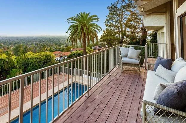 A wooden balcony with seating overlooks a swimming pool, palm tree, and a scenic view of a cityscape and greenery under a clear sky.
