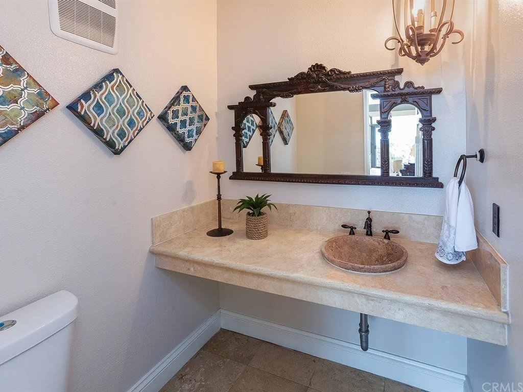 Bathroom with a large ornate mirror, stone countertop and sink, wall-mounted faucet, towel ring, candle, small plant, and decorative wall tiles.