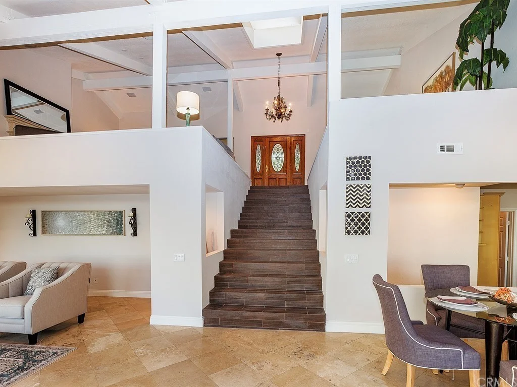 A central staircase with dark steps leads to double wooden doors; living and dining areas with neutral decor are visible on either side.