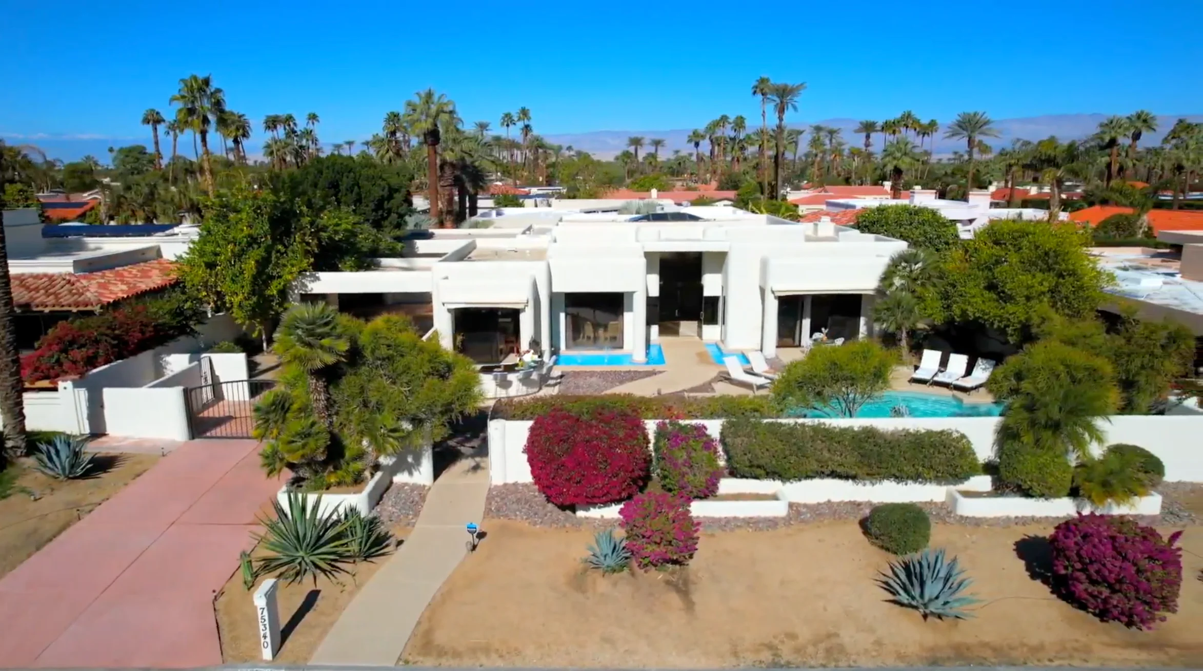 Aerial view of a modern white house with a private pool, surrounded by palm trees, desert landscaping, and mountain views in the background.