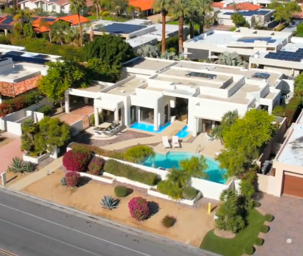 Aerial view of a modern white house with a backyard pool, lounge chairs, landscaping, and surrounding greenery in a suburban neighborhood.
