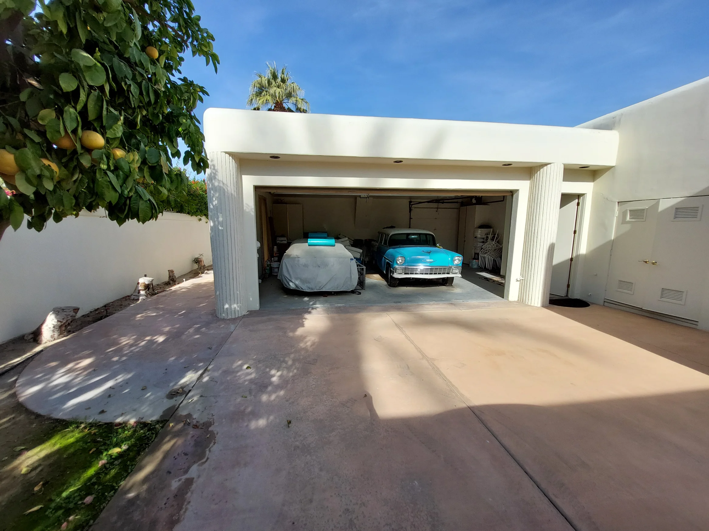A two-car garage with one classic blue car visible and another car covered, next to a tree with yellow fruit and a concrete driveway under a clear sky.