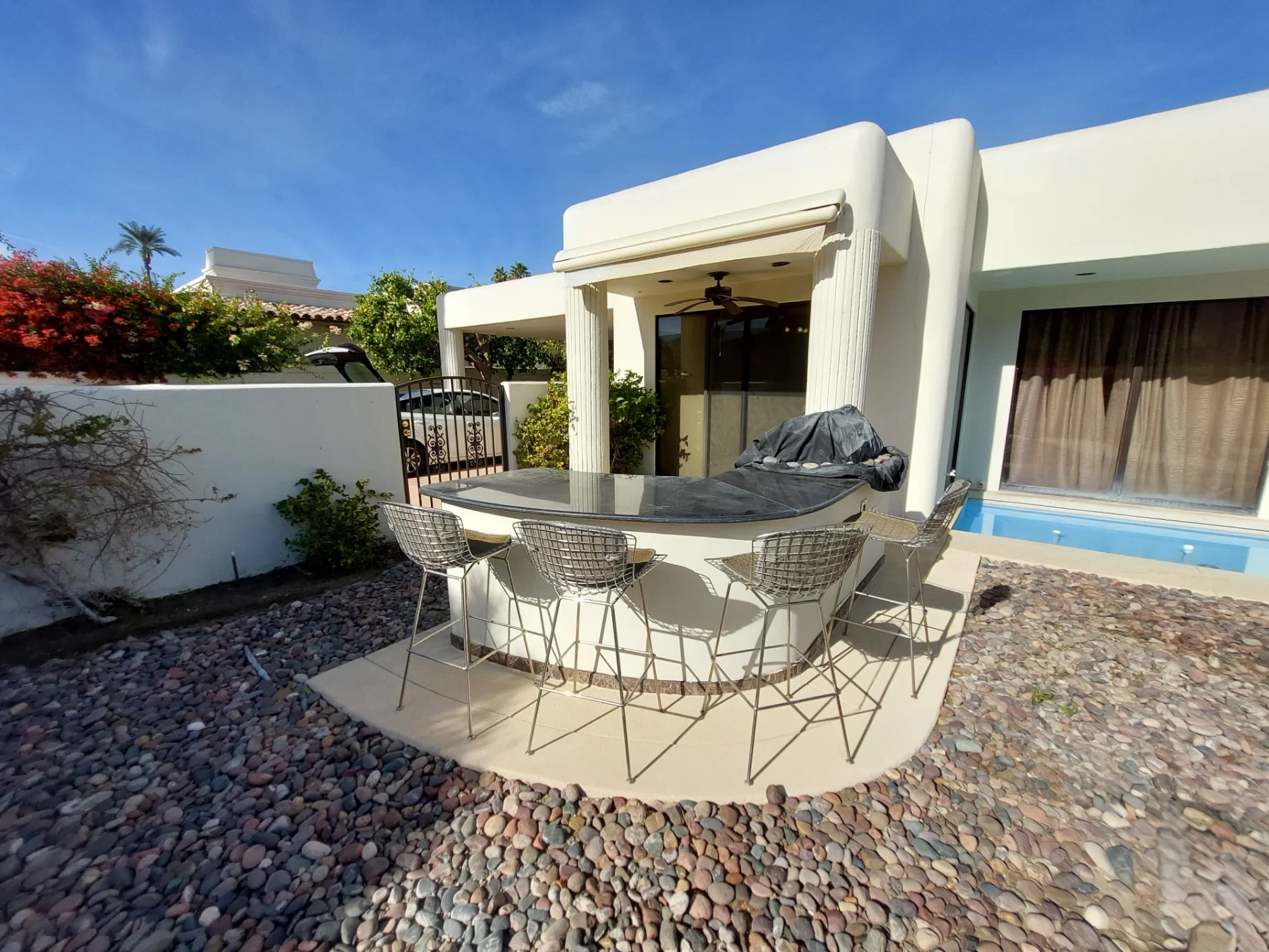 Outdoor patio area with a curved stone bar, four wire chairs, rocky ground covering, and a covered grill next to a white stucco house with large windows and curtains.