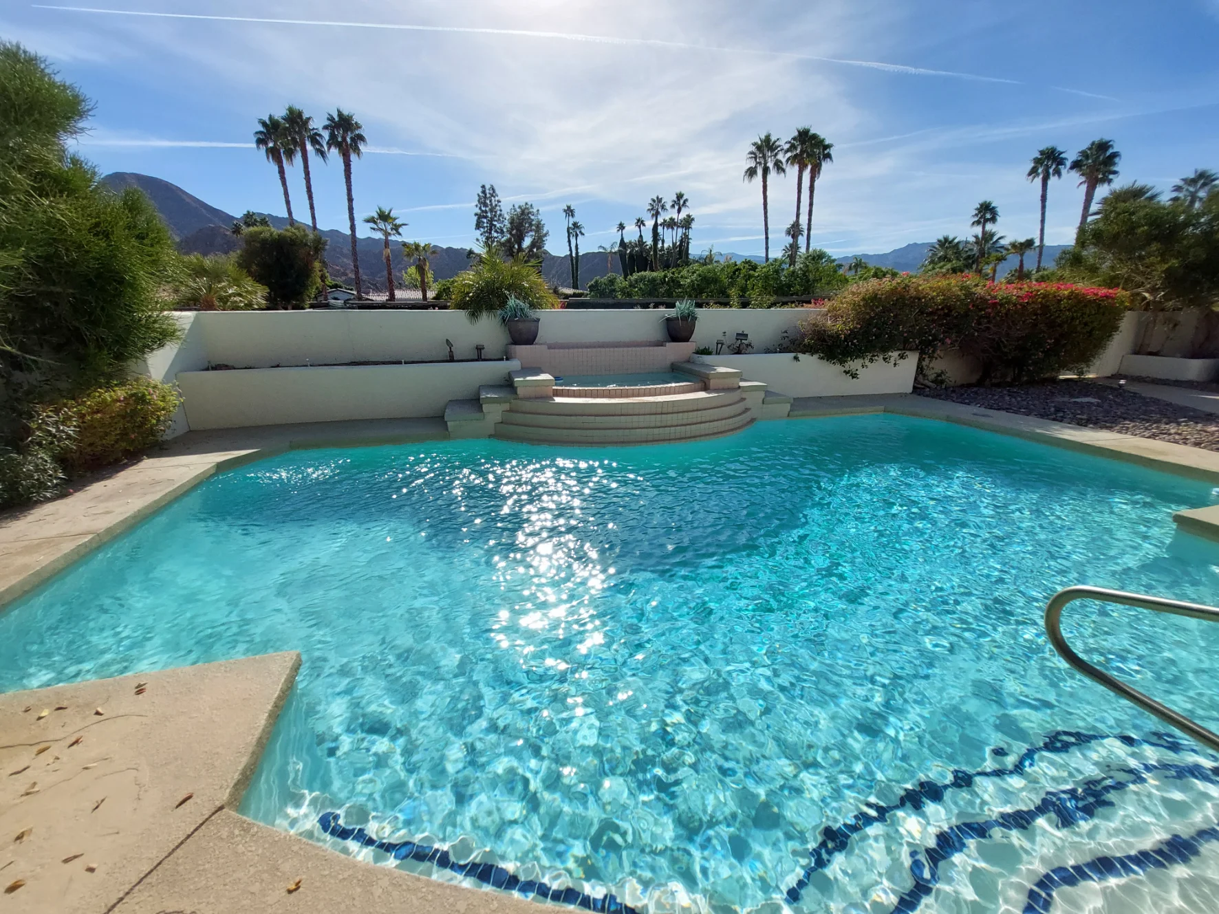A clear swimming pool with steps leading up to a raised platform, surrounded by palm trees and mountains under a sunny sky.