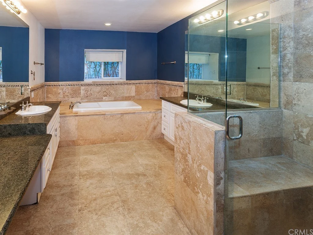 Modern bathroom with tan marble floors and walls, a glass-enclosed shower, built-in bathtub, dual sinks, and blue painted upper walls.
