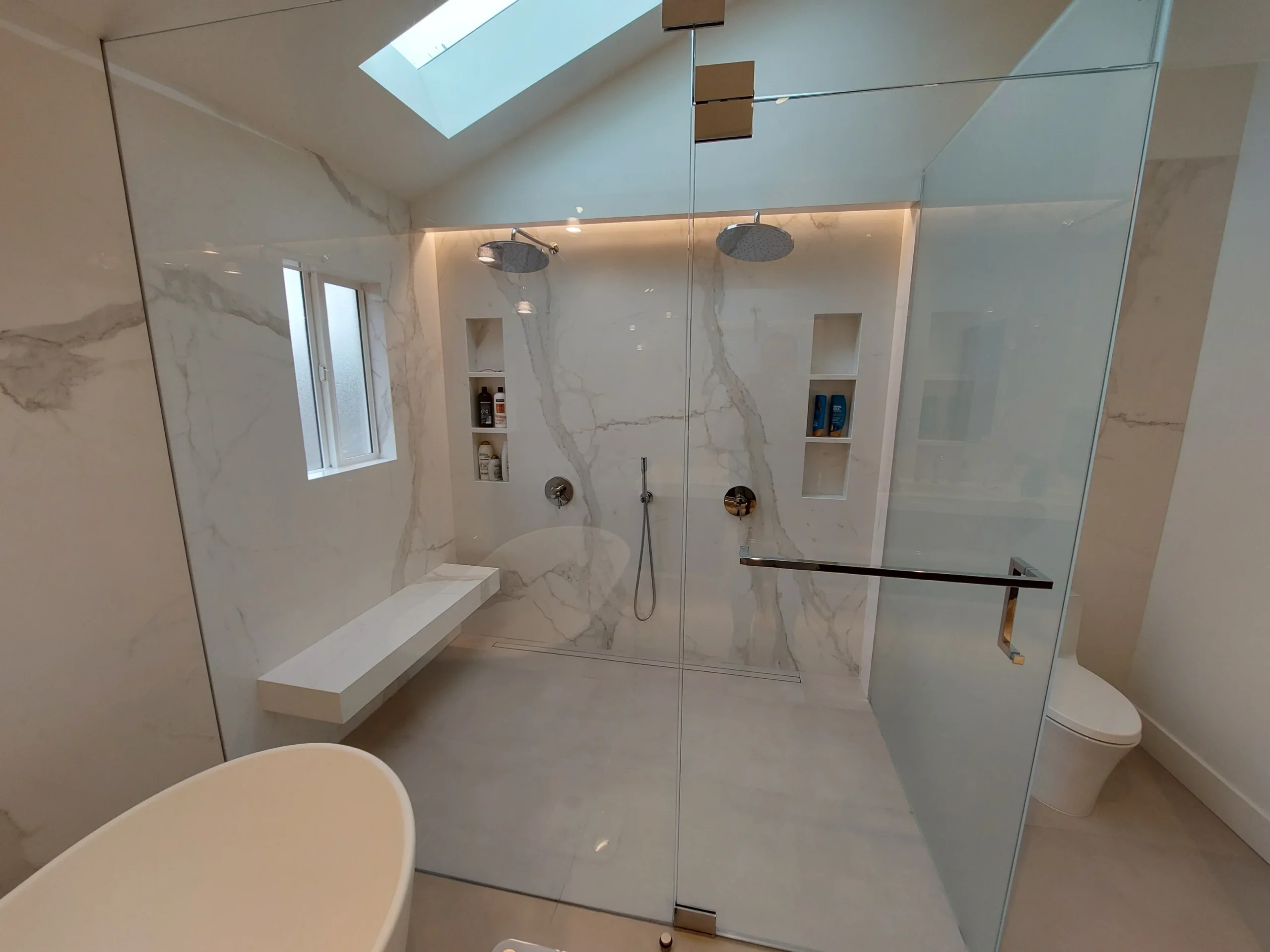 Modern bathroom with a glass-enclosed walk-in shower, dual rain showerheads, built-in shelves, a bench, white walls, and a skylight above.