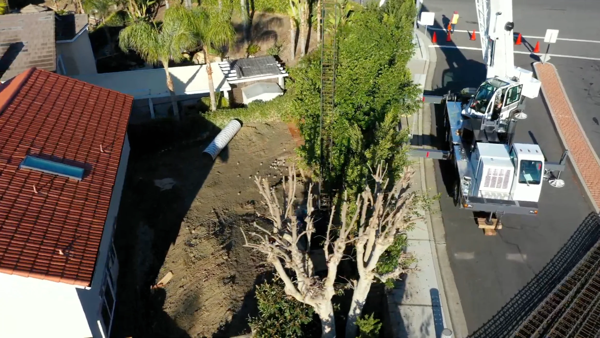 Aerial view of a residential street with a trimmed tree, construction materials, and a work truck parked near orange traffic cones.