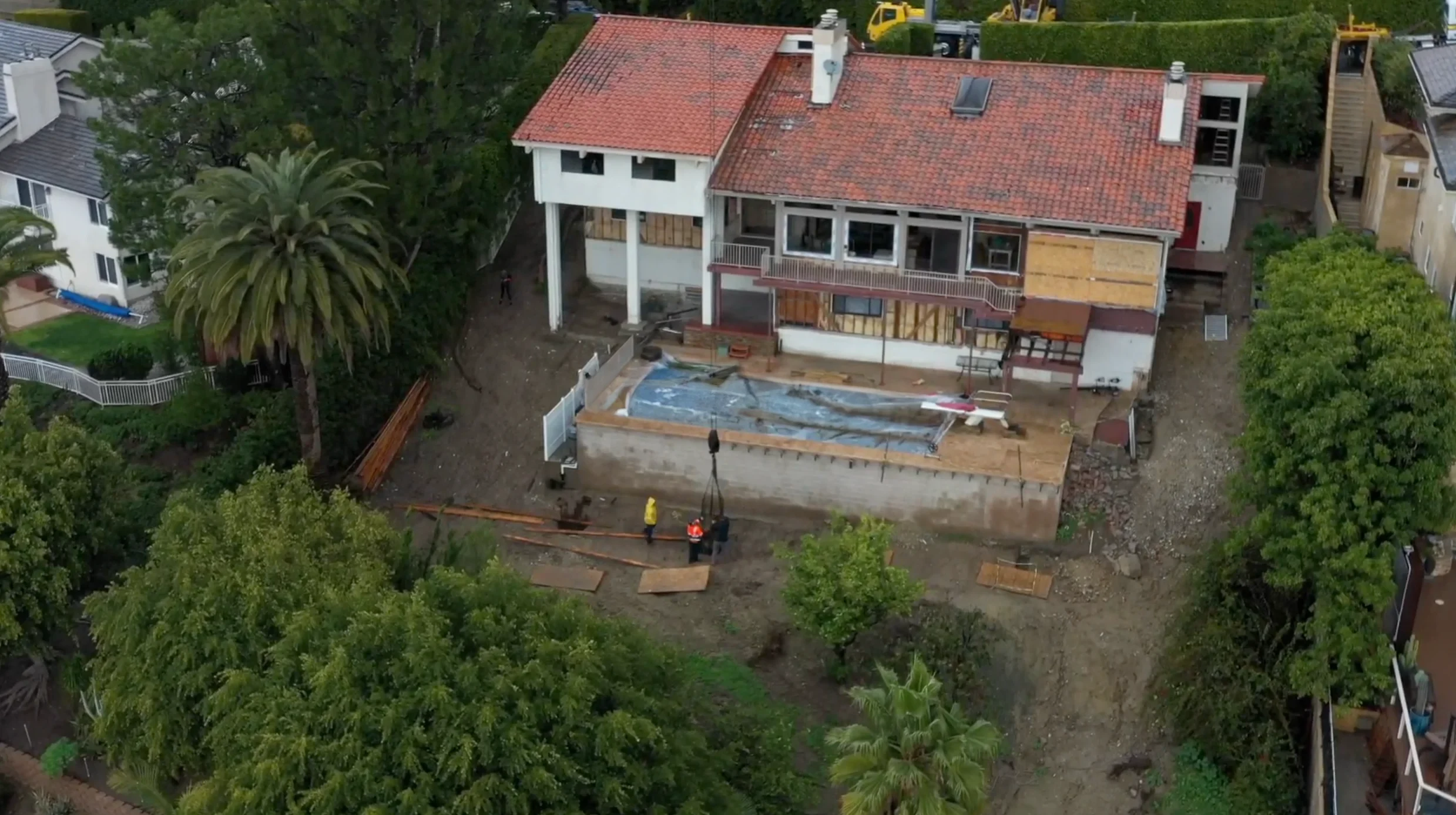 Aerial view of a house under renovation, with exposed walls, construction materials scattered, and an empty pool in the backyard, surrounded by trees.