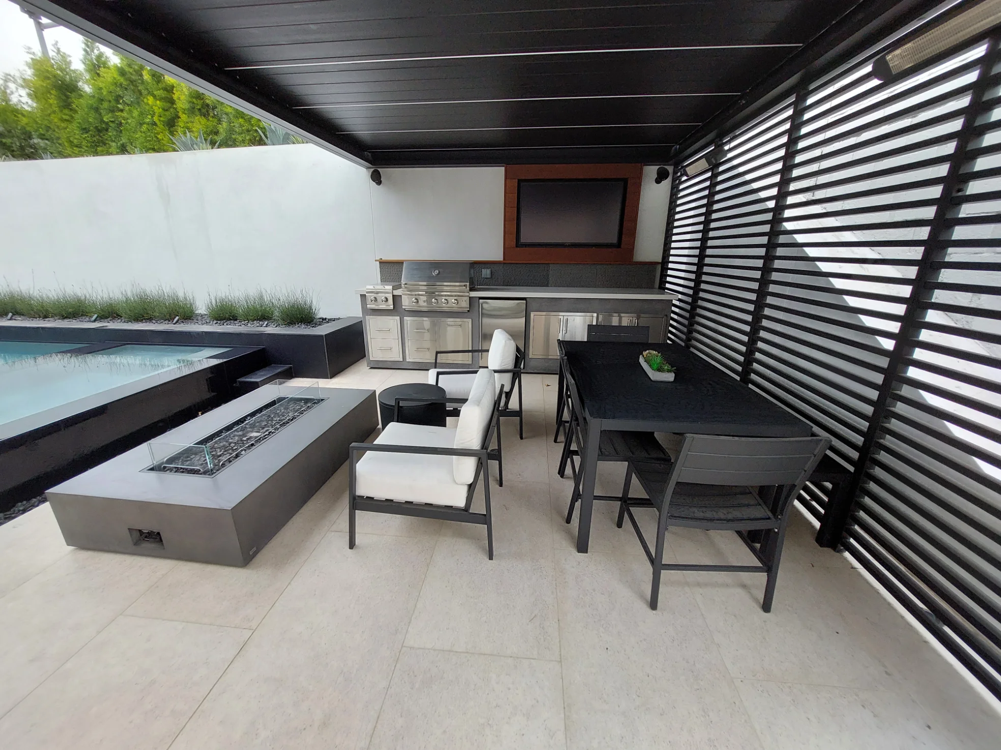 Covered outdoor patio with a grill, television, dining table, lounge chairs, and fire pit next to a pool.
