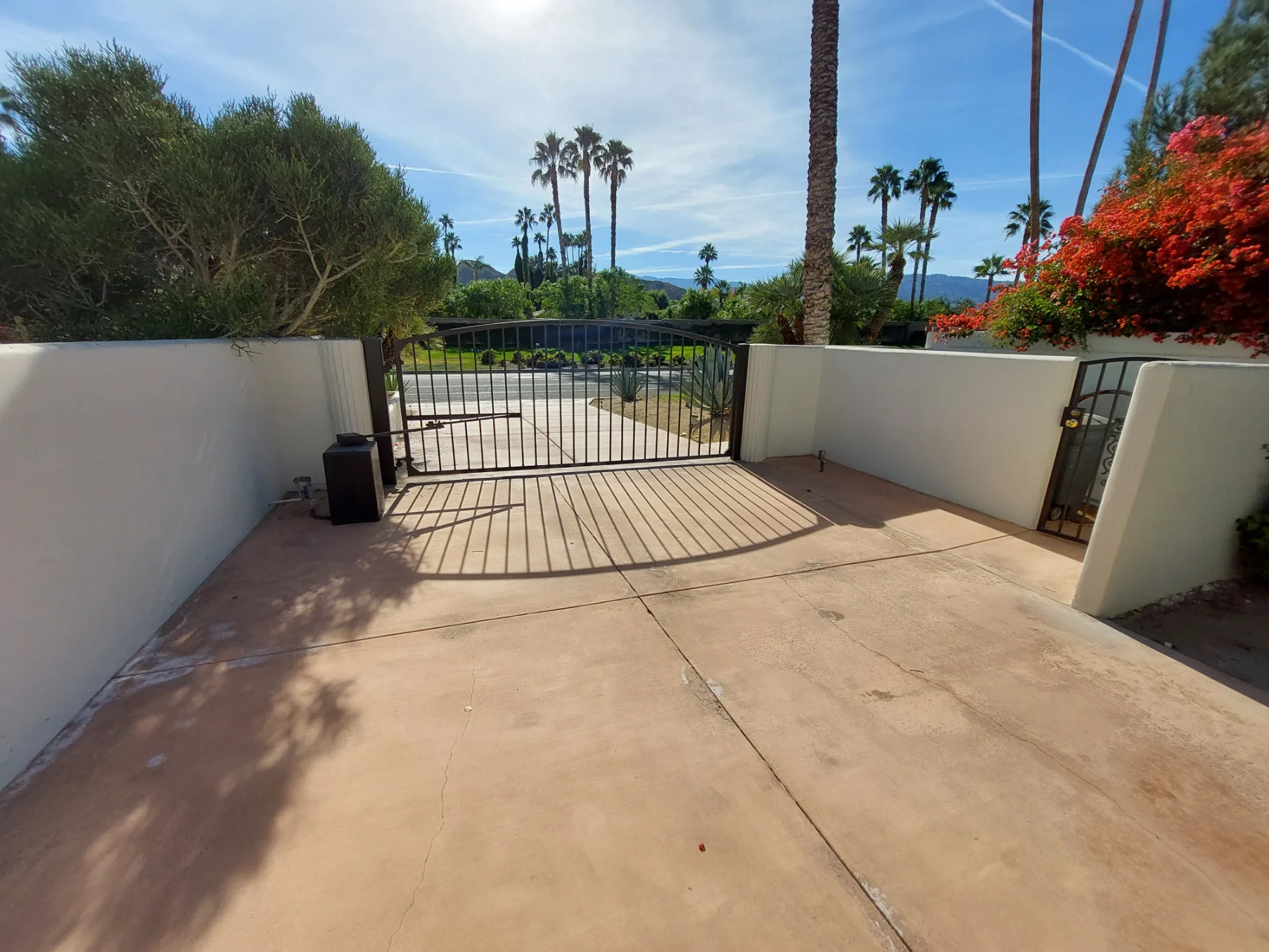 A black metal gate separates a concrete driveway from the street, with palm trees and landscaping visible beyond the gate under a sunny sky.