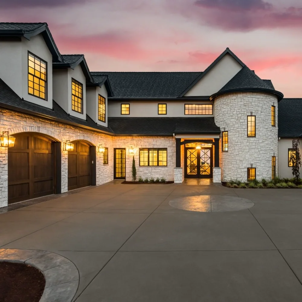 Large modern house by a custom home builder features stone and stucco exterior, three-car garage, round turret, lit windows, and a wide driveway at sunset.