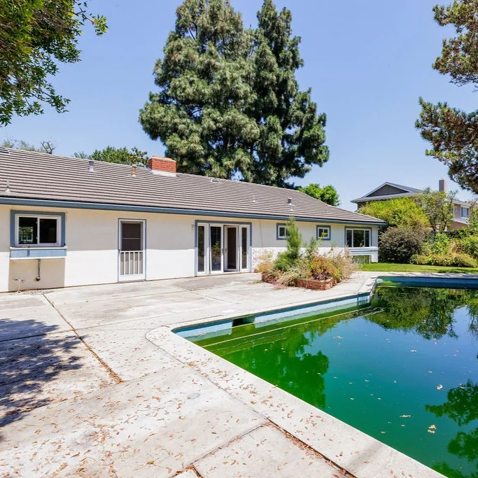 Single-story house with a gray roof and white walls, next to a patio and a rectangular pool with green water, surrounded by trees and shrubs.