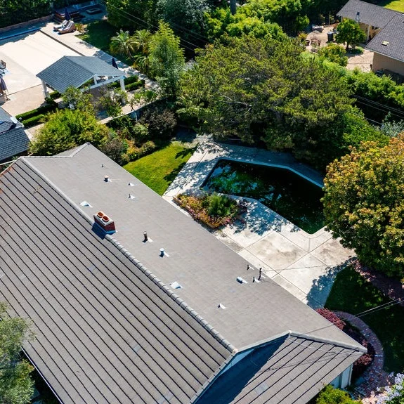 Aerial view of a house with a gray roof, backyard pool, surrounding trees, and neighboring homes visible.