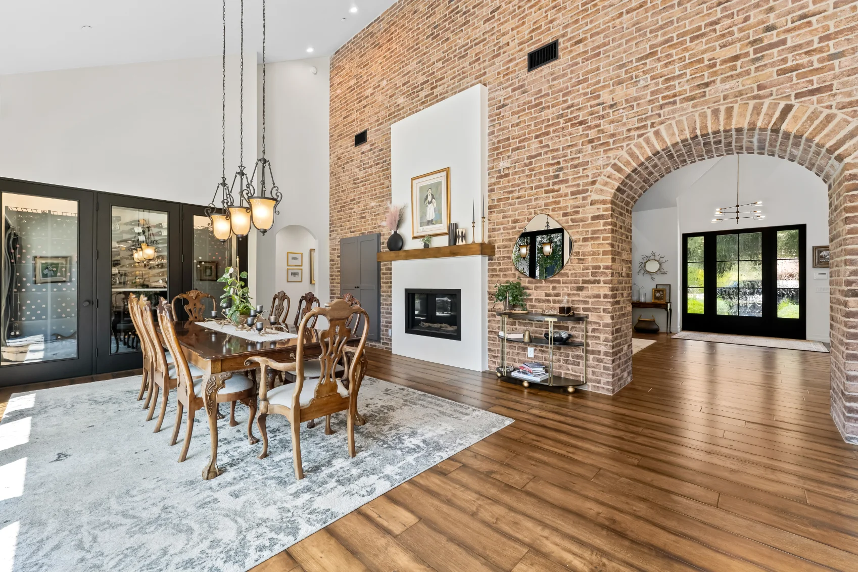 Spacious dining room with a long wooden table, brick accent wall, fireplace, arched doorway, and large windows letting in natural light.