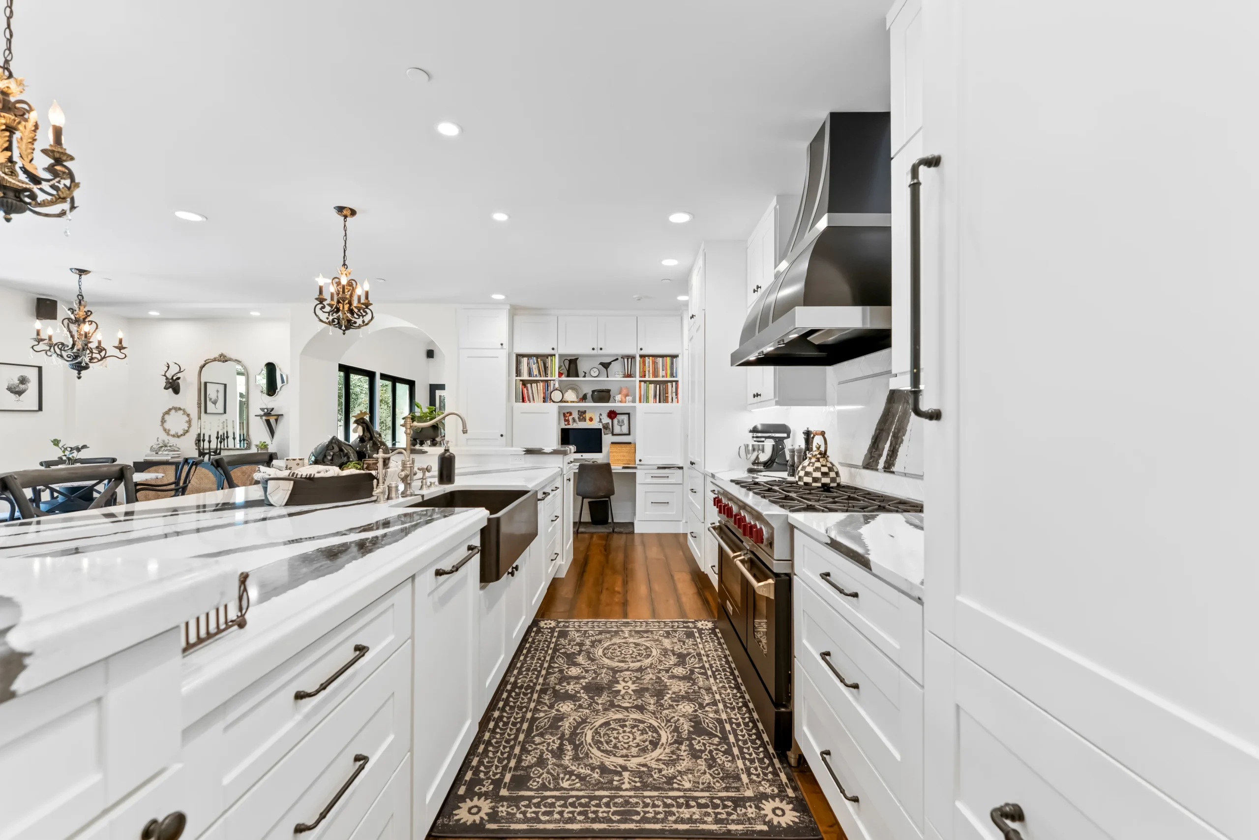 Modern white kitchen with marble countertops, stainless steel appliances, a patterned rug, and a built-in desk area in the background.